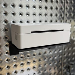 May include: A white label printer on a black shelf, mounted on a silver metal pegboard. The printer has a rectangular shape with a slot for paper output. The pegboard has a diamond pattern.