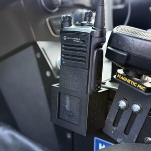 May include: A black Motorola APX 6000 two-way radio mounted in a vehicle. The radio is in a black holder with a small antenna. A "MAGNETIC MIC" label is visible on a nearby component.