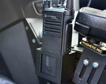 May include: A black Motorola APX 6000 two-way radio mounted in a vehicle. The radio is in a black holder, with an antenna extending upwards. A "MAGNETIC MIC" label is visible nearby.