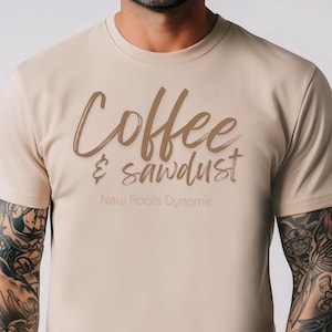 May include: A beige t-shirt with the words "Coffee & sawdust" in brown script. Below the text is "New Roots Dynamic" in a smaller, lighter font. The shirt has short sleeves.