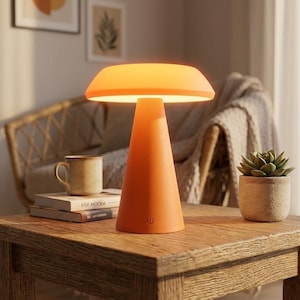 May include: Orange cone-shaped table lamp with a rounded top emitting a warm glow. The lamp sits on a wooden table, next to a mug and a small potted succulent. The lamp has a power button on the base.