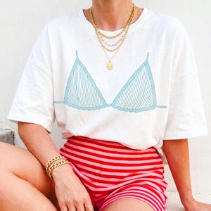 Pode incluir: Uma t-shirt branca de manga curta com um gráfico de linhas em azul-petróleo representando a parte superior de um biquíni no peito. A peça é combinada com calções de riscas horizontais vermelhas e cor-de-rosa, colares e pulseira dourados.