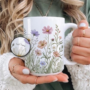 May include: A white ceramic mug featuring a colorful botanical illustration of various wildflowers in shades of pink, purple, and green. The design includes the text "Fuck Trump" integrated into the floral pattern, visible on the main body and a circular inset.