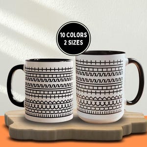 May include: Two white ceramic mugs with black handles and interior. The mugs feature a repeating black geometric pattern. A black circle above the mugs reads "10 COLORS 2 SIZES". The mugs are on a wooden cutting board.