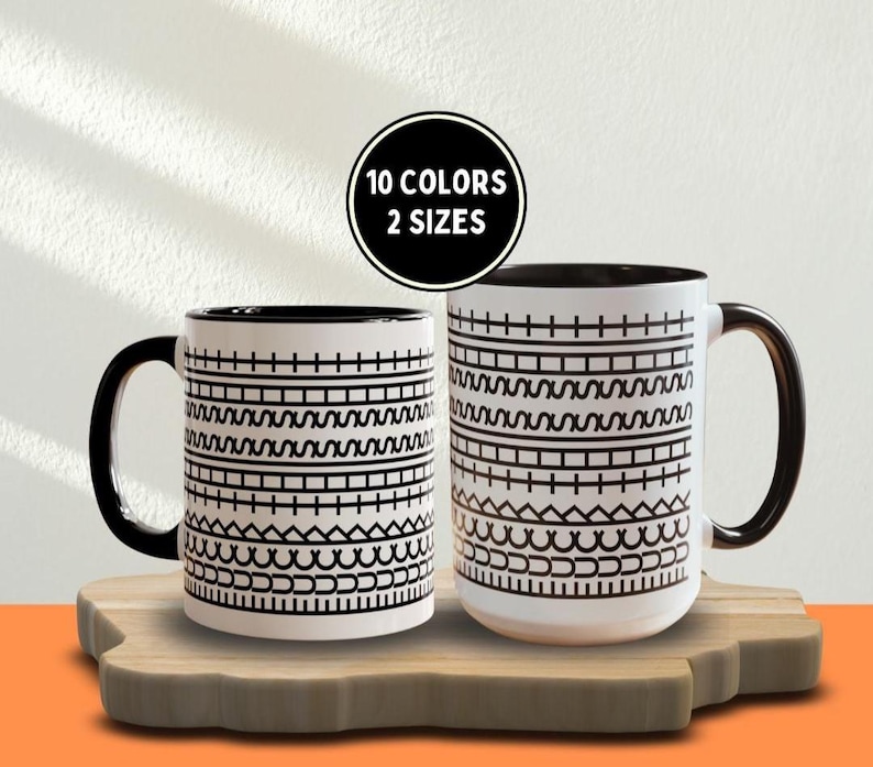 May include: Two white ceramic mugs with black handles and interior. The mugs feature a repeating black geometric pattern. A black circle above the mugs reads "10 COLORS 2 SIZES". The mugs are on a wooden cutting board.