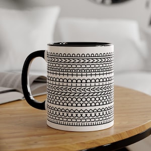 May include: White ceramic mug with a black handle and rim, adorned with a repeating black geometric pattern. The mug sits on a wooden table, showcasing its design. A stylish and functional addition to any kitchen or office.