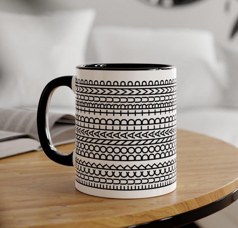 May include: White ceramic mug with a black handle and rim, adorned with a repeating black geometric pattern. The mug sits on a wooden table, showcasing its design. A stylish and functional addition to any kitchen or office.