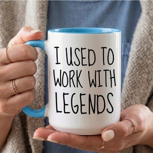 May include: A white ceramic mug with a light blue handle and interior rim. The mug features the black printed text "I USED TO WORK WITH LEGENDS" in a simple sans-serif font centered on the front surface of the cylindrical vessel.