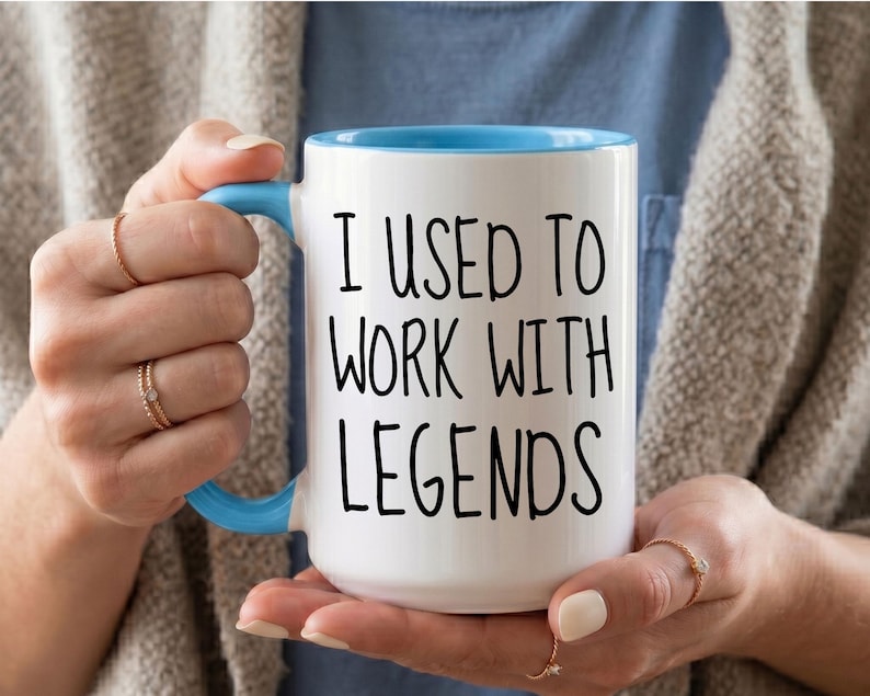 May include: A white ceramic mug with a light blue handle and interior rim. The mug features the black printed text "I USED TO WORK WITH LEGENDS" in a simple sans-serif font centered on the front surface of the cylindrical vessel.