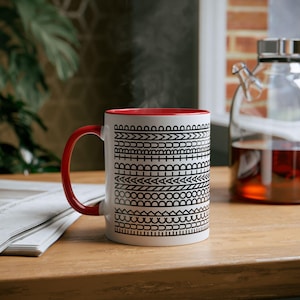 May include: A white ceramic mug with a red interior and handle. The mug features a black geometric pattern around the exterior. Steam rises from the mug, suggesting a hot beverage. The mug sits on a wooden surface.