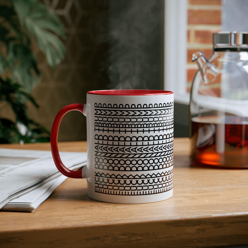 May include: A white ceramic mug with a red interior and handle. The mug features a black geometric pattern around the exterior. Steam rises from the mug, suggesting a hot beverage. The mug sits on a wooden surface.