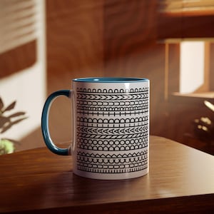 May include: White ceramic mug with a blue interior and handle. The mug features a repeating black geometric pattern around the exterior. The mug is sitting on a wooden surface.