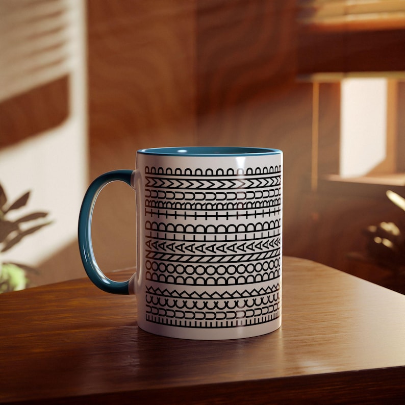 May include: White ceramic mug with a blue interior and handle. The mug features a repeating black geometric pattern around the exterior. The mug is sitting on a wooden surface.