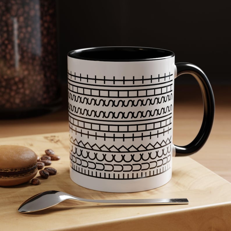 May include: White ceramic mug with a black interior and handle. The mug features a repeating black geometric pattern. A spoon, a macaron, and coffee beans are on a wooden surface.