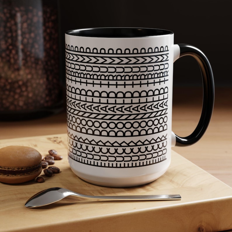 May include: White ceramic mug with a black interior and handle. The mug features a repeating black geometric pattern around the exterior. A spoon and a macaron are on a wooden surface next to the mug.