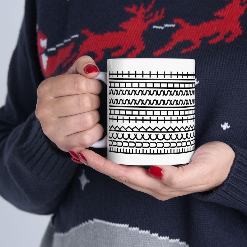 May include: White ceramic mug featuring a black geometric pattern of horizontal lines, zigzags, and other shapes. The mug has a white handle and is held by a person wearing a dark blue sweater.