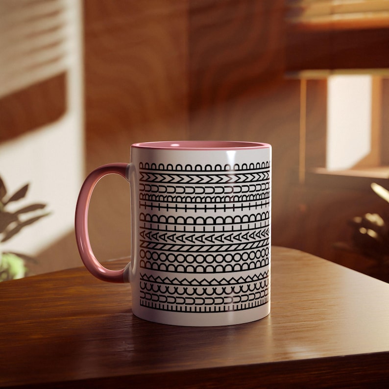 May include: A white ceramic mug with a pink interior and handle. The mug features a repeating black geometric pattern around the exterior. The mug is sitting on a wooden surface.