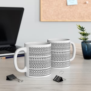May include: Two white ceramic mugs with black geometric patterns. The patterns consist of various shapes and lines, including arrows, circles, and zigzags. The mugs have a standard handle and are placed on a light-colored wooden surface.