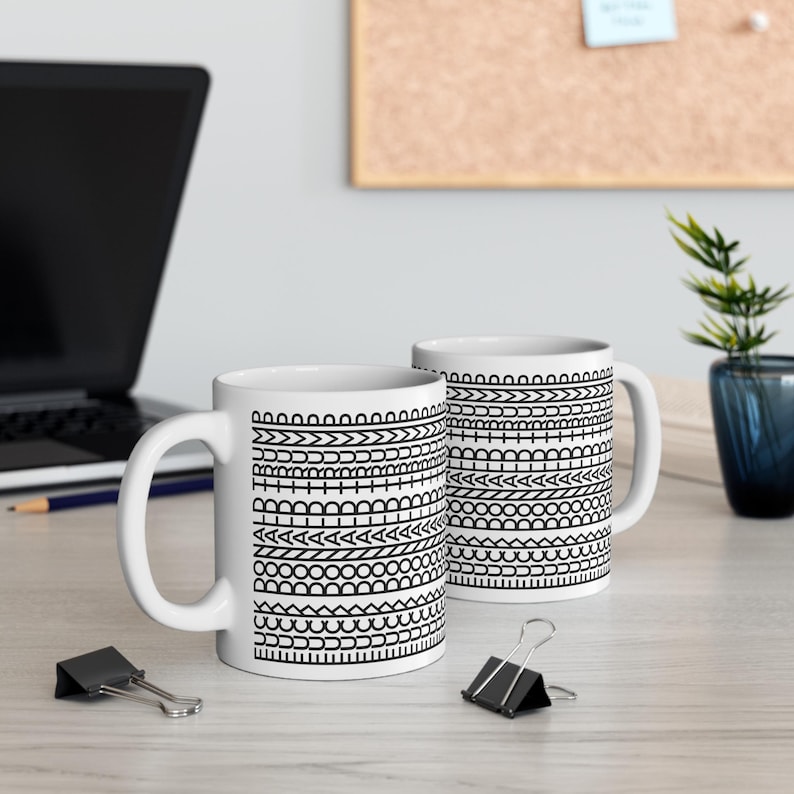 May include: Two white ceramic mugs with black geometric patterns. The patterns consist of various shapes and lines, including arrows, circles, and zigzags. The mugs have a standard handle and are placed on a light-colored wooden surface.