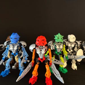 Op de afbeelding: Vijf kleurrijke actiefiguren met verschillende helmen en zilveren wapens. De figuren zijn in blauw, rood, groen, wit en zwart. Ze staan tegen een donkere achtergrond.