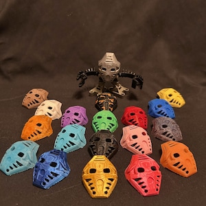 Puede incluir: Una colección de máscaras coloridas impresas en 3D en varios tonos, incluyendo azul, naranja y amarillo, dispuestas alrededor de una figura de acción plateada. Las máscaras tienen diseños intrincados y probablemente son para cosplay o exhibición.
