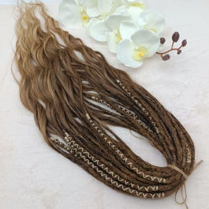 May include: A set of brown and blonde synthetic dreadlocks with white stitching. The dreadlocks are tied together with a brown string.