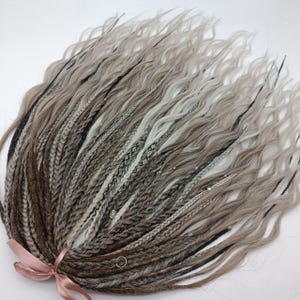 May include: A bundle of gray and brown synthetic dreadlocks with wavy ends. The dreadlocks are tied together with a pink ribbon. The hair extensions feature a mix of colors and textures, including braids and loose strands.