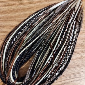 May include: A selection of handmade dreadlocks in shades of brown, black, and cream. The dreads have detailed braiding and decorative elements, such as white stitching and brown ribbons. The arrangement is presented on a wooden surface.