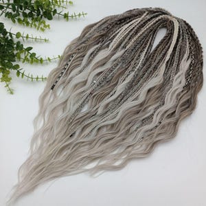 May include: A bundle of synthetic dreadlocks in shades of gray and white. The dreads are a mix of braided and wavy textures, creating a layered effect. The hair extensions are arranged on a white surface, with green foliage in the background.