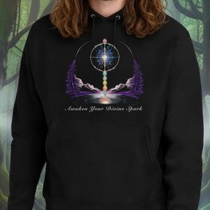May include: Black hoodie with a graphic design featuring a central starburst, stylized trees, and the text "Awaken Your Divine Spark." The design incorporates purple, white, and gold elements. The background is a forest scene.
