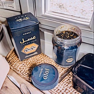 Puede incluir: Una caja y un tarro de miel azul oscuro con el texto "Honey Jojo" en letras doradas. El tarro está abierto, revelando la miel en su interior. También se ve una tapa azul oscuro con el texto "Jojo".