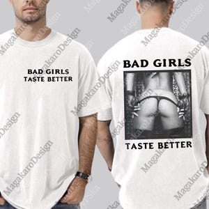 Puede incluir: Camiseta blanca con el texto "BAD GIRLS TASTE BETTER" en la parte delantera. La parte trasera de la camiseta presenta una imagen en blanco y negro de la parte inferior del cuerpo de una persona, con el mismo texto debajo.