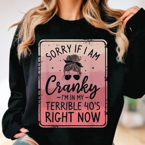 Puede incluir: Sudadera negra con un gráfico rosa y blanco. El gráfico dice "SORRY IF I AM Cranky - I'M IN MY - TERRIBLE 40'S RIGHT NOW" con la cara de una mujer con gafas de sol y un moño.