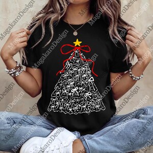 Può includere: T-shirt nero con un design ad albero di Natale fatto di fiocchi bianchi. L'albero è sormontato da una stella gialla e un nastro rosso. Il testo "FOT" è stampato su ogni fiocco. Un capo d'abbigliamento festivo.