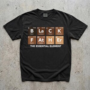 Op de afbeelding: Zwart t-shirt met een periodiek systeem ontwerp dat "BLACK FATHER" spelt. De tekst "THE ESSENTIAL ELEMENT" staat onder het ontwerp.