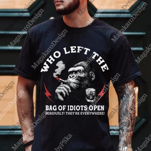May include: Black t-shirt featuring a graphic of a gorilla smoking a cigar with a glass of whiskey. The text reads "WHO LEFT THE BAG OF IDIOTS OPEN SERIOUSLY! THEY'RE EVERYWHERE!" in white font.