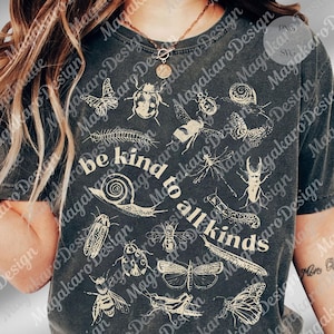 May include: Charcoal grey t-shirt with white insect drawings, such as butterflies and beetles. The phrase "be kind to all kinds" is printed across the front in a vintage style. A nature-inspired design, perfect for casual wear.