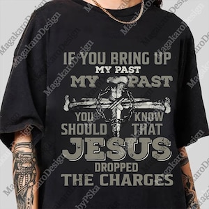 May include: Black t-shirt with a graphic design featuring a cross and the text "IF YOU BRING UP MY PAST, YOU SHOULD KNOW THAT JESUS DROPPED THE CHARGES." The shirt has a crew neck and short sleeves. A gold chain is visible.