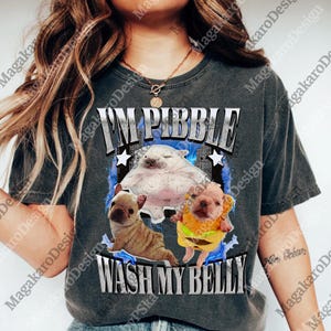 Pode incluir: T-shirt cinza escuro com um gráfico que diz "I'M PIBBLE WASH MY BELLY". O design apresenta três animais de desenho animado, um branco grande e dois filhotes. O gráfico tem um efeito de raio azul e estrelas brancas.