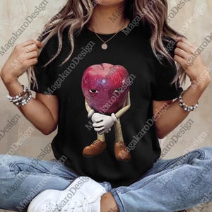May include: Black t-shirt featuring a cartoon apple character with a face, white gloves, and brown shoes. The apple is red with a purple hue. The shirt is worn by a person wearing blue jeans and white sneakers.