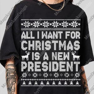 以下が含まれることがあります： 黒いTシャツに白いニットのデザイン。テキスト「ALL I WANT FOR CHRISTMAS IS A NEW PRESIDENT」が、雪の結晶とトナカイのグラフィックとともに、お祝いのフォントで表示されています。上部には金のチェーンが見えます。