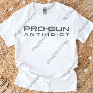 May include: A white t-shirt with the text "PRO-GUN ANTI IDIOT" printed in grey. The shirt has a crew neck and is made of a soft material. The shirt is displayed on a neutral background.