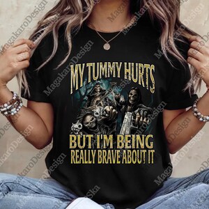 May include: Black t-shirt with the text "MY TUMMY HURTS BUT I'M BEING REALLY BRAVE ABOUT IT" and a graphic of grim reapers holding guns. The design is in gold and white.