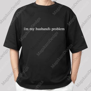 May include: Black t-shirt with the text "i'm my husband's problem" in white. The shirt has short sleeves and a relaxed fit. The text is centered on the front of the shirt.