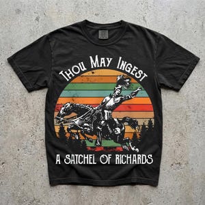 May include: A black short-sleeved t-shirt featuring a graphic of a knight on horseback against a sunset background with horizontal stripes in orange, yellow, and green. The text reads "THOU MAY INGEST A SATCHEL OF RICHARDS" in a white serif font.