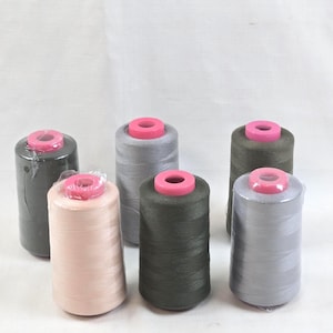 May include: A collection of six cylindrical spools of sewing thread in various shades of gray, charcoal, and light pink. Each spool features a bright pink plastic cap at the top and is wrapped in clear protective plastic film for storage.