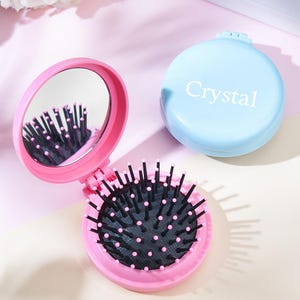 Peut inclure: Un miroir compact rose avec une brosse à cheveux noire à l'intérieur. Le miroir est ouvert et la brosse est visible. Le compact porte l'inscription "Crystal" en blanc sur le côté.