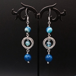 May include: A pair of dangle earrings with blue and silver accents. Each earring features a silver hook, a blue bead, a silver ring with a smaller blue bead, and a larger blue bead at the bottom. The earrings are displayed on a black stand.
