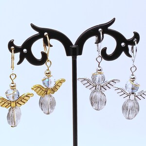 May include: A pair of angel earrings with gold-colored wings and a clear, faceted body. The earrings are displayed on a black stand. The wings are detailed with etched lines, and the head is a clear, iridescent bead. The earrings have a lever-back closure.
