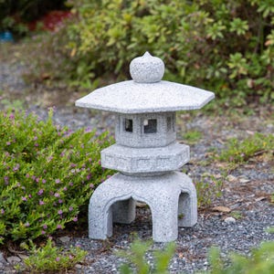 Puede incluir: Una linterna de jardín japonesa de piedra gris con un diseño de varios niveles. La linterna presenta un techo hexagonal, ventanas cuadradas y una parte superior redondeada. Se apoya sobre una base curva, rodeada de vegetación y pequeñas rocas.